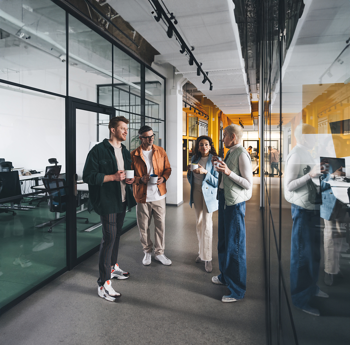 Group of multiracial team members wearing casual clothes standing in corridor of modern office and talking to each other with cups in hands during coffee break